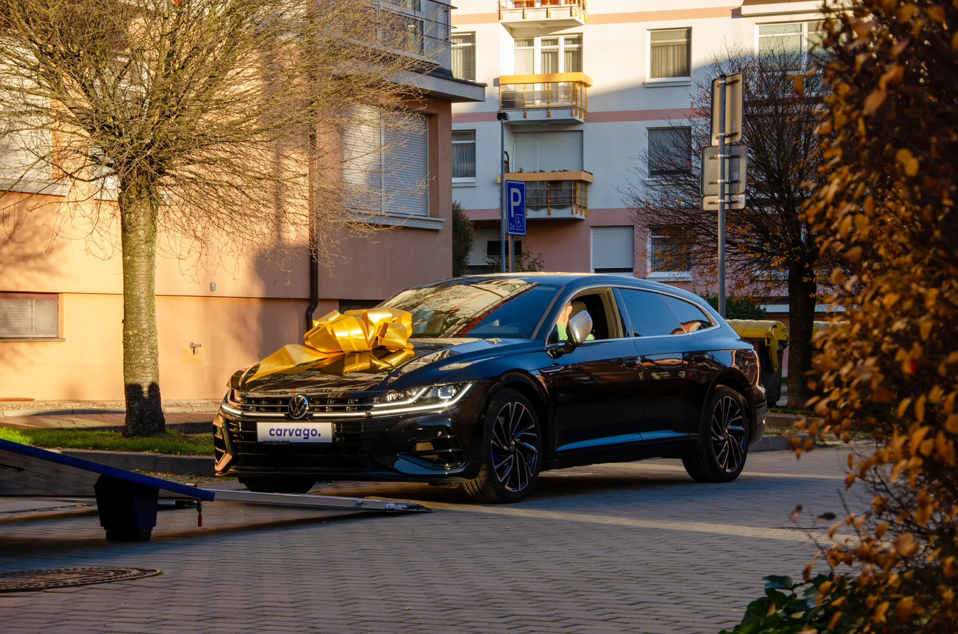 Black Volkswagen Arteon with a yellow ribbon bow on the hood, parked in a residential area during golden hour with a transporter partially visible on the left