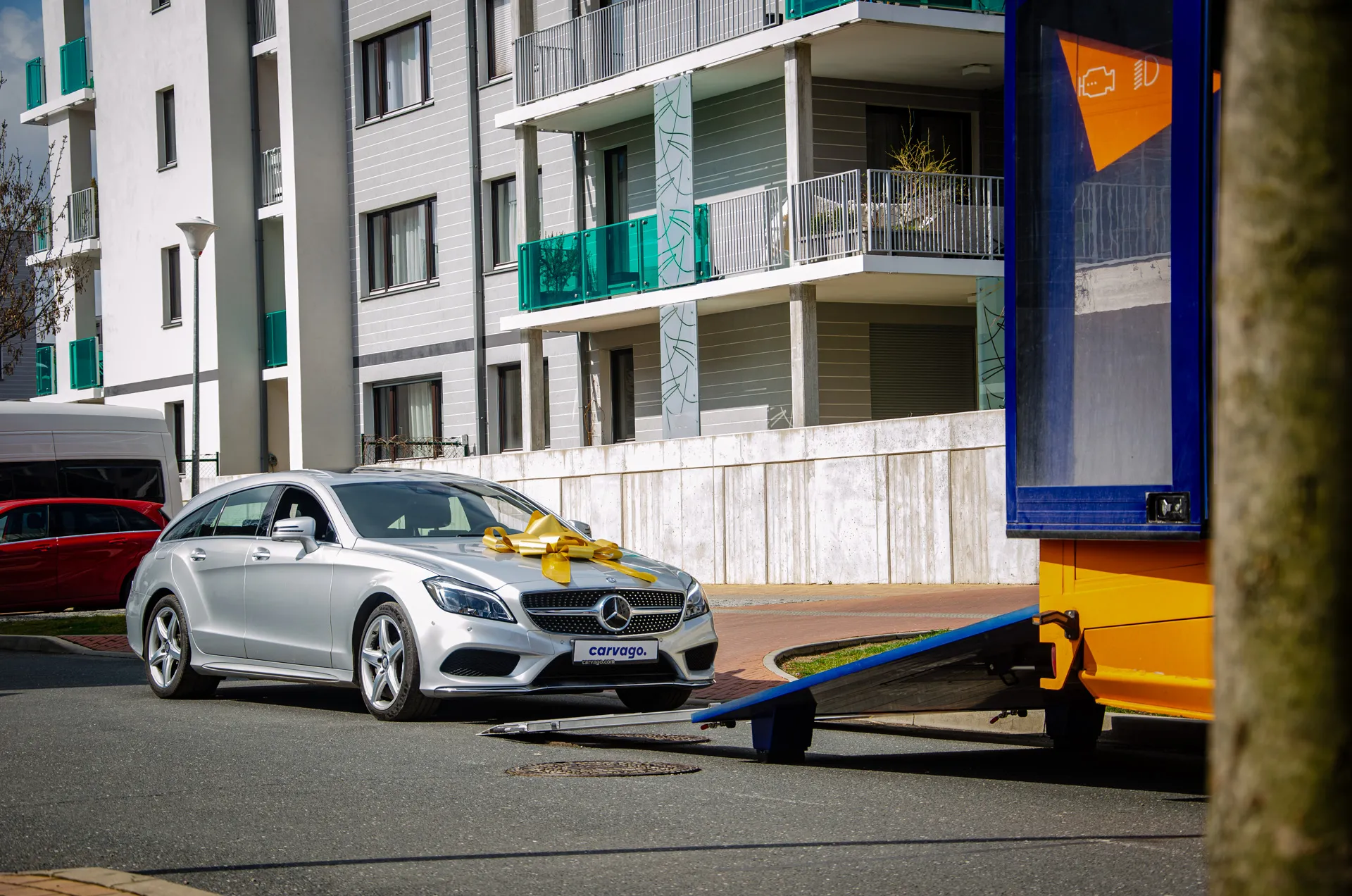 Silver Mercedes-Benz wagon with a yellow ribbon bow on the hood loading onto a car transporter ramp, with panel buildings in the background