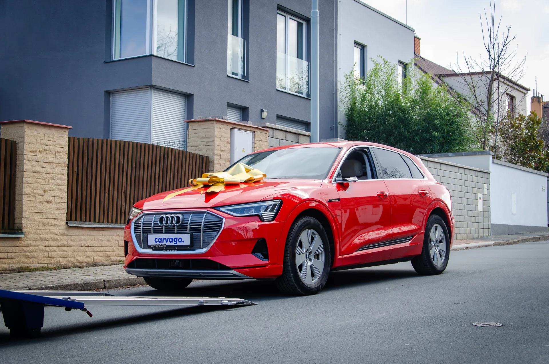 Red Audi e-tron SUV with a yellow ribbon bow on the hood parked by a metal fence in a modern urban street