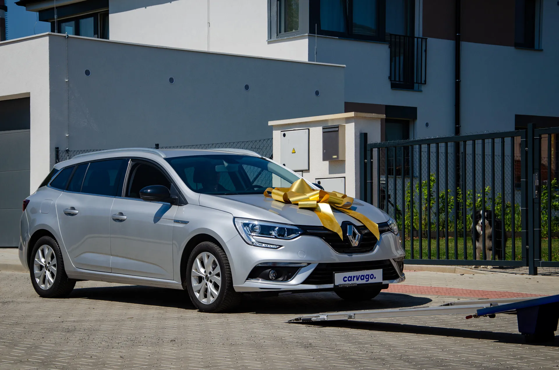 Silver Renault Megane wagon with a yellow ribbon bow on the hood parked in front of a modern grey house, with a transporter partially in frame